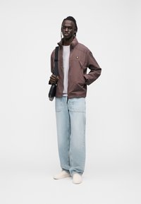 Giacca marrone leggera con colletto alto, T-shirt bianca sotto, jeans a gamba larga azzurri chiari e scarpe beige slip-on. Borsa a tracolla nera.