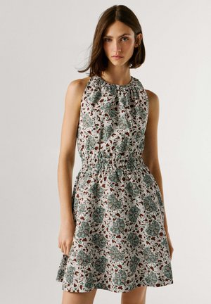 Jeune femme portant une robe sans manches avec un col et une taille froncés, arborant un motif floral vert et marron sur fond blanc.