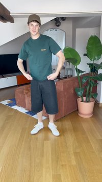 Zelené bavlněné tričko, černé denimové šortky a bílé tenisky. Vnitřní prostředí s dřevěnou podlahou a rostlinou v květináči na pozadí.