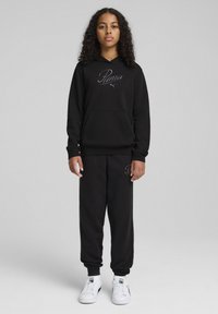 Sweat-shirt à capuche noire avec logo brodé, associé à un pantalon de survêtement avec poignets élastiques. Dispose d'une grande poche frontale et de baskets blanches.