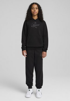 Sweat-shirt à capuche noire avec logo brodé, associé à un pantalon de survêtement avec poignets élastiques. Dispose d'une grande poche frontale et de baskets blanches.