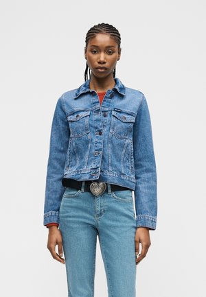 Νεαρή γυναίκα με πλεξουδωτά μαλλιά φοράει ένα μπλε τζιν μπουφάν, ανοιχτόχρωμο μπλε τζιν παντελόνι, κόκκινη μπλούζα και μαύρη ζώνη με καρδιοειδή αγκράφα.