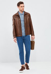 Giacca di pelle marrone, polo azzurro chiaro, jeans di denim azzurro chiaro, sneakers marroni, con una borsa marrone. Outfit semplice e informale.