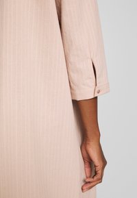 Bras droit d'une personne portant une chemise rose clair à manches longues avec un bouton au poignet sur fond gris.