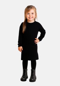 Schwarzes, geripptes Strickkleid mit langen Ärmeln, kombiniert mit schwarzen Leggings und klobigen schwarzen Stiefeletten. Einfaches Design, figurschmeichelnde Passform und strukturierter Stoff.