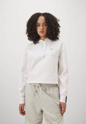 White hooded sweatshirt with a cropped design, featuring a small logo on the chest. Paired with light beige drawstring pants.