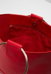 Rød syntetisk håndtaske med en glat yderside og guldfarvede metalringe. Indersiden har et stoffor og en lynlåslomme.