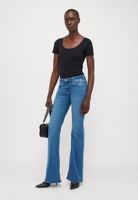 Svart åtsittande t-shirt, blå utsvängda jeans, mjukt denimmaterial och svart handväska med silverdetaljer. Modellen står i en neutral miljö.