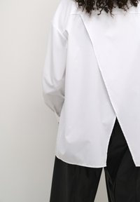 Hvid skjorte med lange ærmer, der har et delt rygdesign, glat tekstur og knapmanchetter, parret med sorte skinnende bukser.