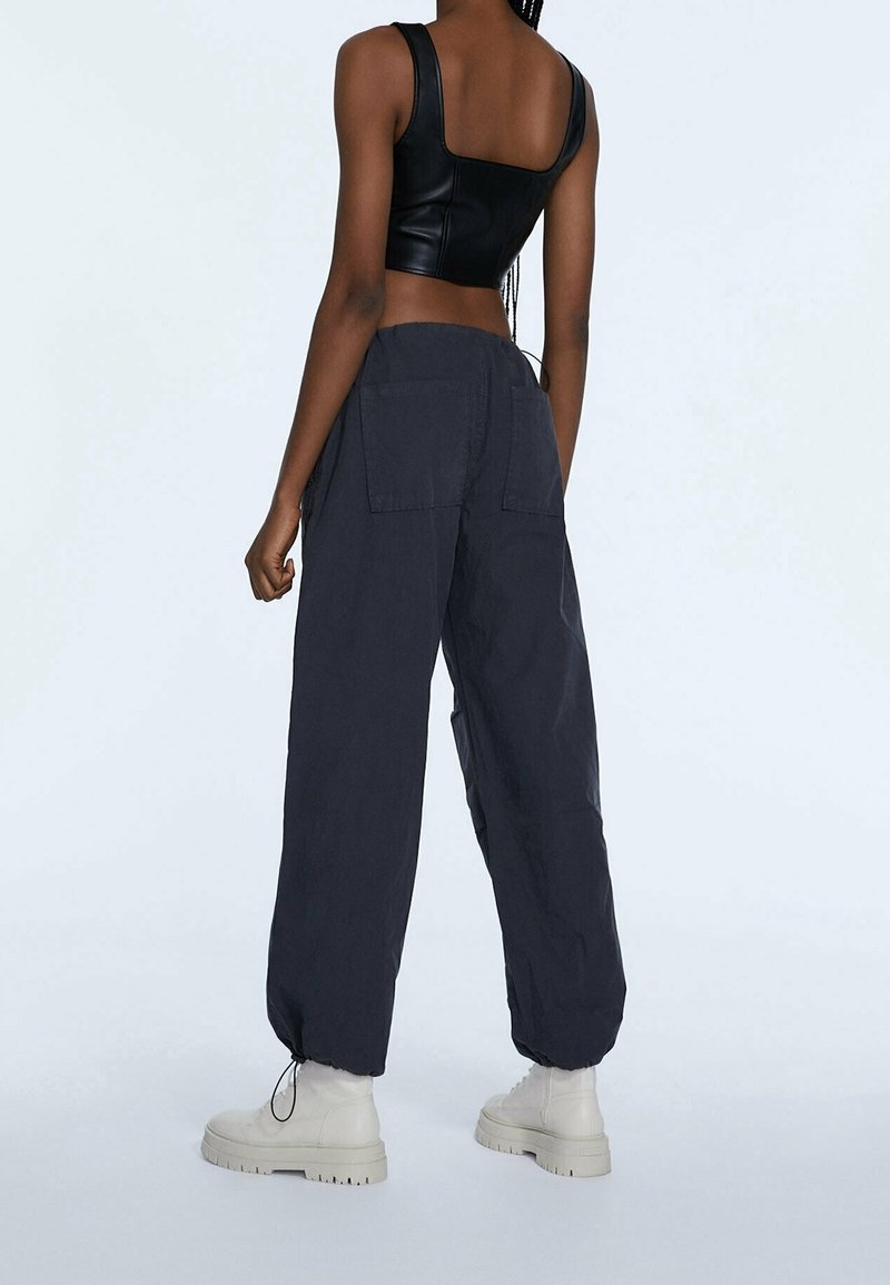 Top en cuir noir court avec des bretelles larges, associé à un pantalon cargo gris foncé ample avec poches latérales et poignets élastiques.