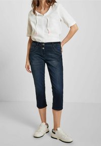 Tmavě modré denimové capri kalhoty se dvěma předními kapsami, zakřiveným pasem a kontrastním prošíváním. Spárované s bílým topem s krátkým rukávem a teniskami.