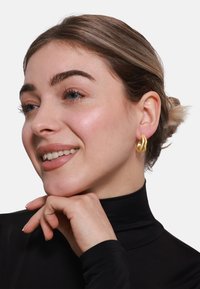 Gullfargede hoop-øreringer med en tvunnet design, med en glatt overflate og en subtil glans, båret i en models øre mot en svart topp.