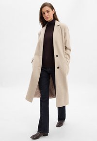 Beige Wollmantel mit Reverskragen, doppelt geknöpftem Verschluss und seitlichen Taschen, getragen über einem dunklen Rollkragenpullover und blauen Jeans.
