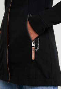 Main dans la poche d'une veste noire avec une fermeture éclair argentée visible et un curseur en cuir marron, texte brodé à côté de la poche, personne portant un jean bleu.