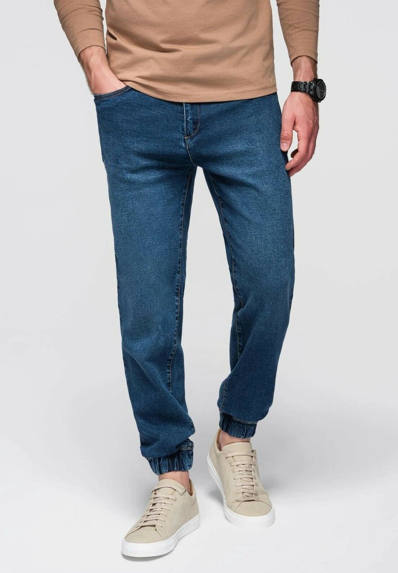 Joggers de mezclilla azul con puños elásticos, combinados con zapatillas beige. La tela es suave, con un diseño clásico de cinco bolsillos y un corte relajado.