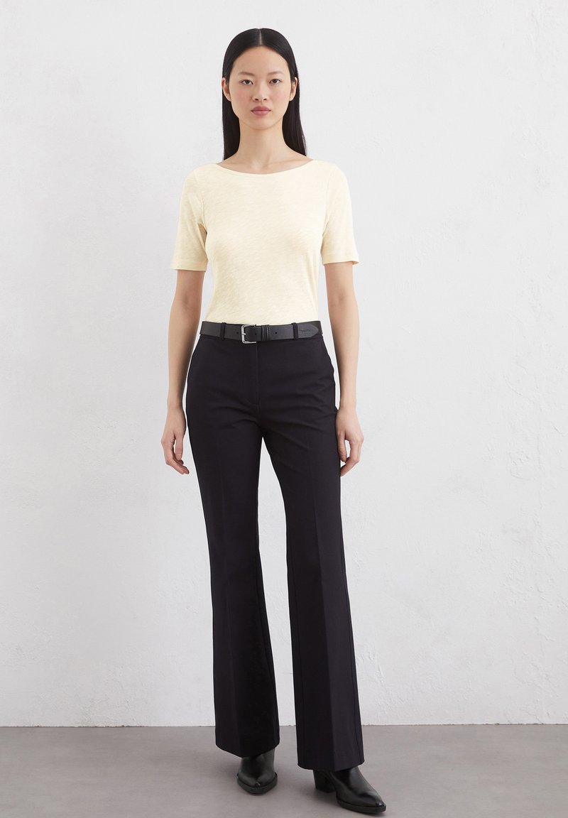 Kurzärmliges, hellgelbes Oberteil mit Boot-Ausschnitt, kombiniert mit schwarzen Schlaghosen und schwarzen, knöchelhohen Stiefeln mit Absatz. Einfaches Design, glatte Textur.