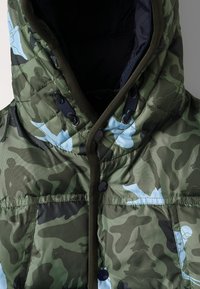Chaqueta de plumas de camuflaje verde con textura acolchada, cuello alto y acentos azules. Presenta botones a presión y cordón en la capucha.