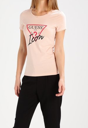 Mujer con camiseta de manga corta de color rosa claro con texto negro "Guess Icon" y pantalones negros sobre un fondo blanco liso.