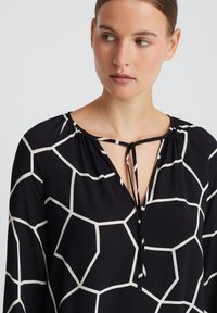 Zwarte blouse met wit hexagonaal patroon, voorzien van een v-hals met striksluiting en geraffineerde details bij de nek. Zachte stof, lange mouwen.