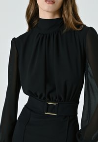 Schwarze Bluse mit hohem Kragen, gerafftem Stoff an den Schultern, durchsichtigen langen Ärmeln und einer taillierten Taille mit einer goldenen rechteckigen Gürtelschnalle.