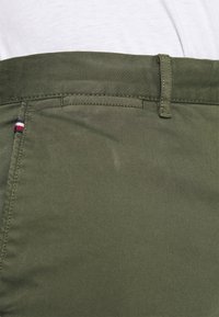 Olijfgroene katoenen broek met een gladde textuur, voorzien van een gestikte tailleband en een klein rood, wit en blauw logo op de zak.