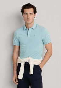 Mladý muž v světle modrém polo tričku s malým logem, tmavě modrých kalhotách a bílém svetru uvázaném kolem pasu, stojící před jednoduchým pozadím.