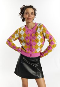 Pletený kardigan s argyle vzorem v růžov�é, žluté a bílé barvě, s kulatým výstřihem, předními knoflíky a žebrovanými manžetami. V kombinaci s černou koženou sukní.