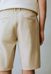 Shorts en coton beige avec deux poches arrière à fermeture boutonnée et une texture lisse. La coupe est décontractée, se terminant au-dessus du genou.