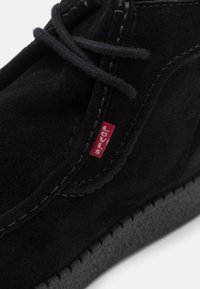 Bottine en daim noir avec lacets plats et semelle en caoutchouc texturée. Présente une petite étiquette rouge avec un détail de logo sur le côté.