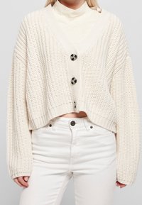 Tjock creamfärgad stickad cardigan med djup V-hals, oversize ärmar och två svarta rundade knappar framtill. Kort modell.