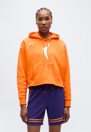 Donna con capelli intrecciati che indossa una felpa corta arancione con un logo di pallacanestro e pantaloncini sportivi viola.