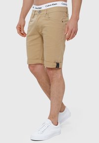Beige bomulds-shorts med en omfoldet kant, der har en synlig knap og lynlås lukning. Taljen viser "Calvin Klein" branding, parret med hvide sneakers.