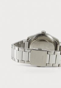 Montre en acier inoxydable avec un bracelet lisse et poli et un fond circulaire. La boucle présente une gravure "GUESS". Design simple sans motifs.