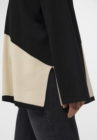 Maglione nero e beige con un design a blocchi di colore diagonali, fessure laterali, tessuto a maglia testurizzato e un orlo nero a contrasto.