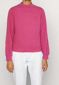 Roze sweatshirt met een hoge kraag, geribde manchetten en zoom. Voorzien van een klein logo op de borst. Gecombineerd met witte broek. Zachte textuur.