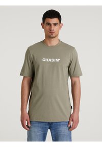 Olíva zöld póló, puha anyagból készült, rövid ujjú és kerek nyakú. Az elején fehér "CHASIN'" felirat található.
