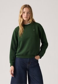 Sweatshirt vert à manches longues, avec des poignets côtelés et un col rond, présentant un petit logo sur la poitrine, associé à un pantalon en denim large de couleur foncée.