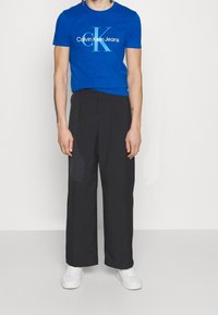 Camiseta de algodón azul con el logo blanco de Calvin Klein Jeans, combinada con pantalones negros holgados y zapatillas blancas. Un conjunto simple y casual.