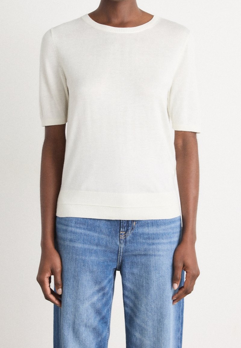 Femme portant un haut en tricot blanc à manches courtes et unicolore, associé à un jean en denim bleu, sur fond blanc.