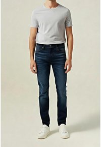 Camiseta de manga corta gris claro combinada con vaqueros ajustados azul oscuro y zapatillas blancas, de pie sobre un fondo neutro.