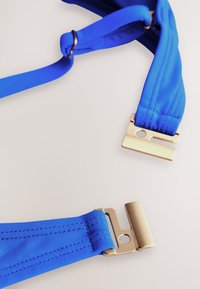 Sangle en tissu bleu avec boucle en métal doré et anneau réglable sur fond clair uni.