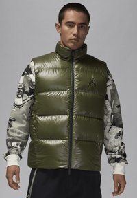 Olivgrön pufferväst med hög krage, med en frontdragkedja och glänsande yta. Modellen bär en mönstrad långärmad skjorta underneath.