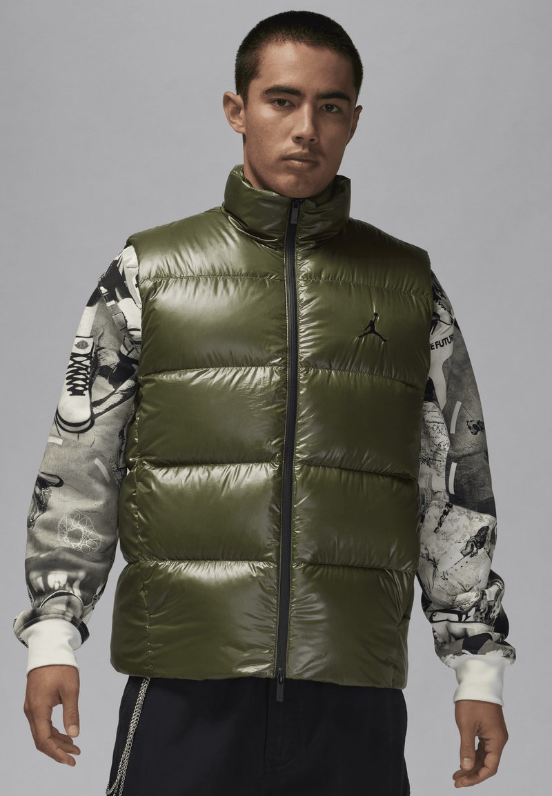 Olivgrön pufferväst med hög krage, med en frontdragkedja och glänsande yta. Modellen bär en mönstrad långärmad skjorta underneath.