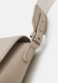 Beige lædertaske med tekstureret overflade, justerbar rem med sølvfarvet hardware og syningsdetaljer. Nærbillede af lukkemekanisme.