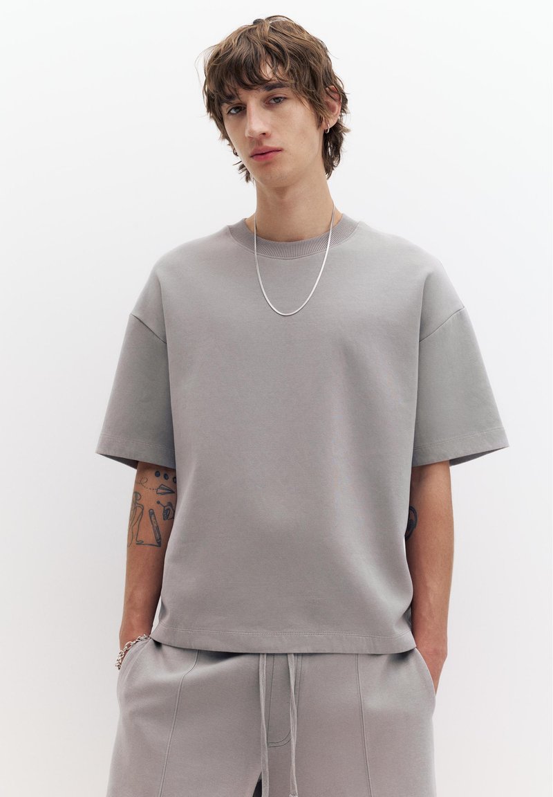 Jeune homme aux cheveux bruns ébouriffés, portant une chemise à manches courtes grise ample et un pantalon assorti, collier en chaîne argentée, tatouages visibles sur les bras.