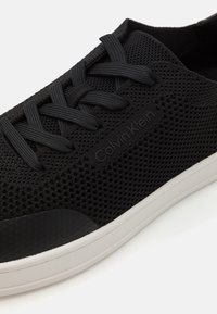 Baskets noires texturées avec une tige en mesh, des lacets noirs et une semelle en caoutchouc blanche. Présente un logo Calvin Klein sur le côté.