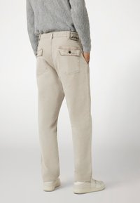Beige katoenen broek met twee achterzakken, een merkembleem bij de tailleband en een taps toelopende fit. Draagt met witte sneakers.
