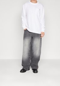 Vit långärmad T-shirt med bröstficka, kombinerad med grå jeans i vida ben med en gradienttoning. Svarta skor fullbordar outfiten.