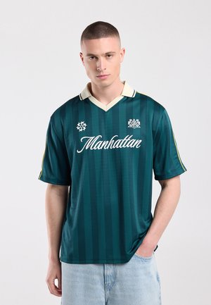 Jonge man met kort haar die een groen gestreept sportshirt draagt met de tekst "Manhattan" en lichtblauwe spijkerbroek tegen een effen achtergrond.