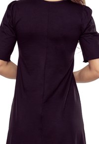 Na zdjęciu przedstawiono plecy osoby w ciemnofioletowej koszuli z krótkimi rękawami, podkreślającej gładki materiał i prosty krój.